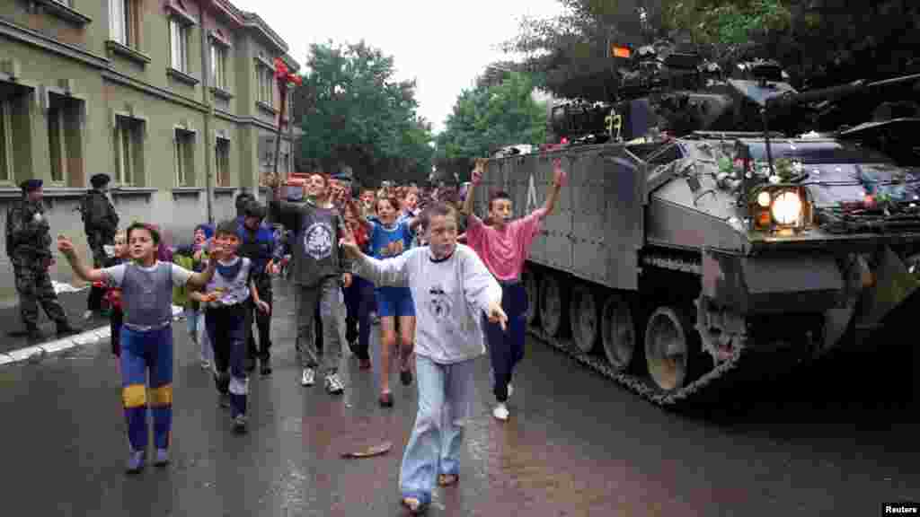 Podujevë 18 qershor 1999: Forcat e KFOR-it arritën në Podujevë disa ditë më vonë se në qytetet tjera të Kosovës. Në këtë fotografi shihen fëmijët duke vrapuar pranë një tanku derisa përshëndesin ushtarët britanikë, të cilët sapo kishin arritur në qytet. &nbsp; 6 qershor 2024: Njëzetepesë vjet pas, në të njëjtin vend objekti komunal mbetet thuajse i njëjtë. Sheshi kryesor është zgjeruar dhe janë ndërtuar objekte të reja. 
