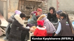 Марал Шаболотова вместе с детьми сидит за столом рядом со снесенным домом. 