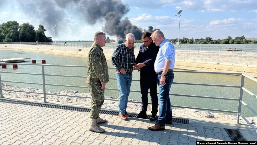 Ministrul Apărării din România, Angel Tîlvăr (al doilea din stânga), a vizitat malul românesc al Dunării, unde au fost găsite resturi de drone rusești, după gafele de comunicare la cel mai înalt nivel din statul român. În spate: incendiul provocat de un atac pe malul ucrainean.