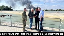 Ministrul Apărării din România, Angel Tîlvăr (al doilea din stânga), a vizitat malul românesc al Dunării, unde au fost găsite resturi de drone rusești, după gafele de comunicare la cel mai înalt nivel din statul român. În spate: incendiul provocat de un atac pe malul ucrainean.