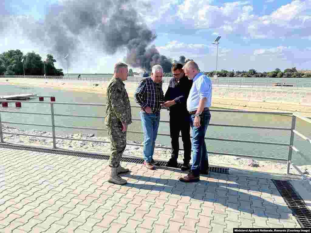 Smoke rises from Ukrainian territory as Romania&rsquo;s Defense Minister Angel Tilvar (in plaid shirt) consults with other men in an image released by the ministry on September 6. In a September 6 Facebook post, Romania&rsquo;s Defense Ministry appeared to contradict the assertions of the president, announcing it had discovered &ldquo;elements that resemble drone remains&rdquo; on Romanian territory just across the border from Ukraine&#39;s Izmayil, which it vowed to investigate.