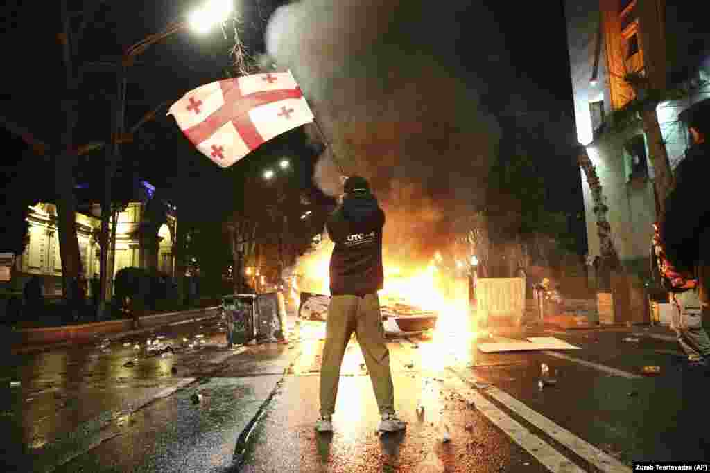 Një burrë valëvit një flamur kombëtar gjeorgjian përpara një barrikade që është duke u djegur gjatë protestave në Tbilisi javën që shkoi kundër një projektligji të diskutueshëm për &quot;agjentët e huaj&quot;.