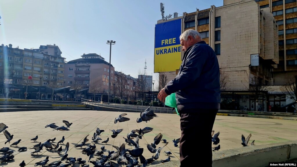 Një burrë duke ushqyer pëllumba në kryeqytetin e Kosovës më 24 shkurt, teksa në prapavijë shihet mesazhi i solidarizimit të Prishtinës me Ukrainën.