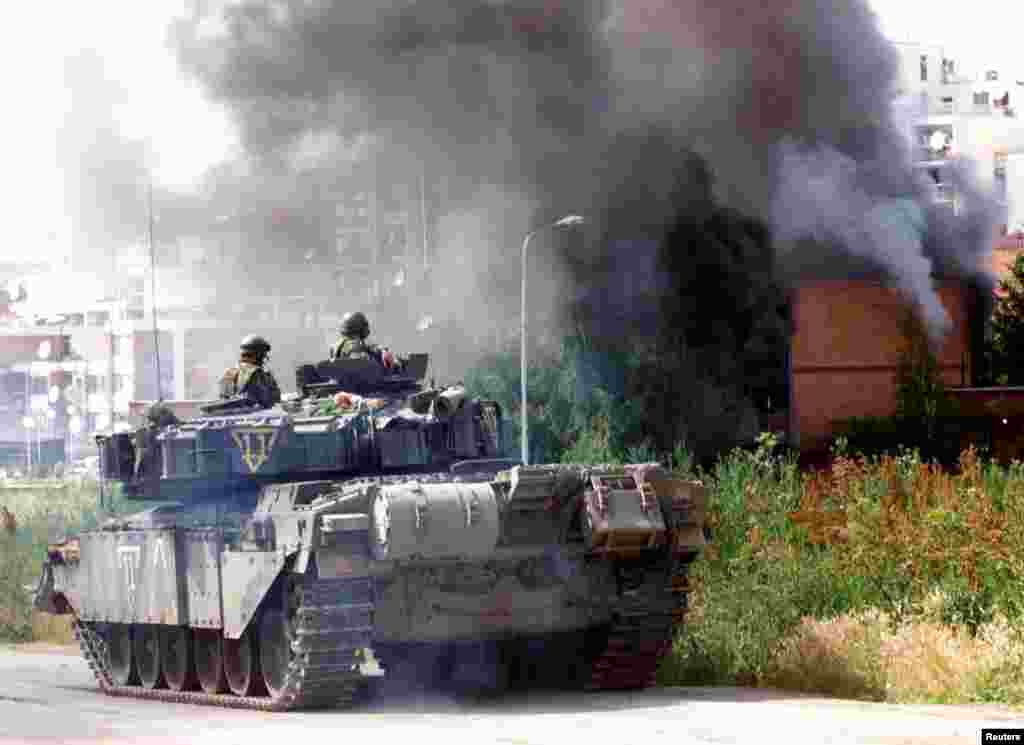 Soldați britanici privesc clădirile care ard în Priștina, 13 iunie 1999, în urma unui atac al unui soldat sârb. O nouă școală și numeroase proiecte imobiliare se află de-a lungul drumului unde odinioară ardeau casele.