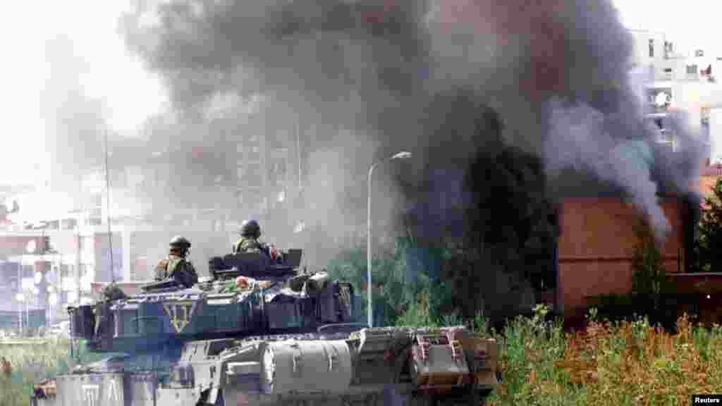 Prishtinë 13 qershor 1999: Ushtarët britanikë derisa shikojnë një objekt banimi kolektiv duke u djegur në Prishtinë. Sipas përshkrimit të fotografisë nga Reuters, kjo fotografi është shkrepur pak kohë pasi trupat britanike ishin sulmuar me armë zjarri nga një pjesëtar i ushtrisë serbe. &nbsp; 5 qershor 2024: Përgjatë viteve shihet se në këtë lokacion është ndërtuar një objekt shkollor, si dhe janë vendosur rrethoja pas zgjerimit të rrugës. Shumë objekte të tjera për banim kolektiv janë ndërtuar në 15 vjetët e fundit. 