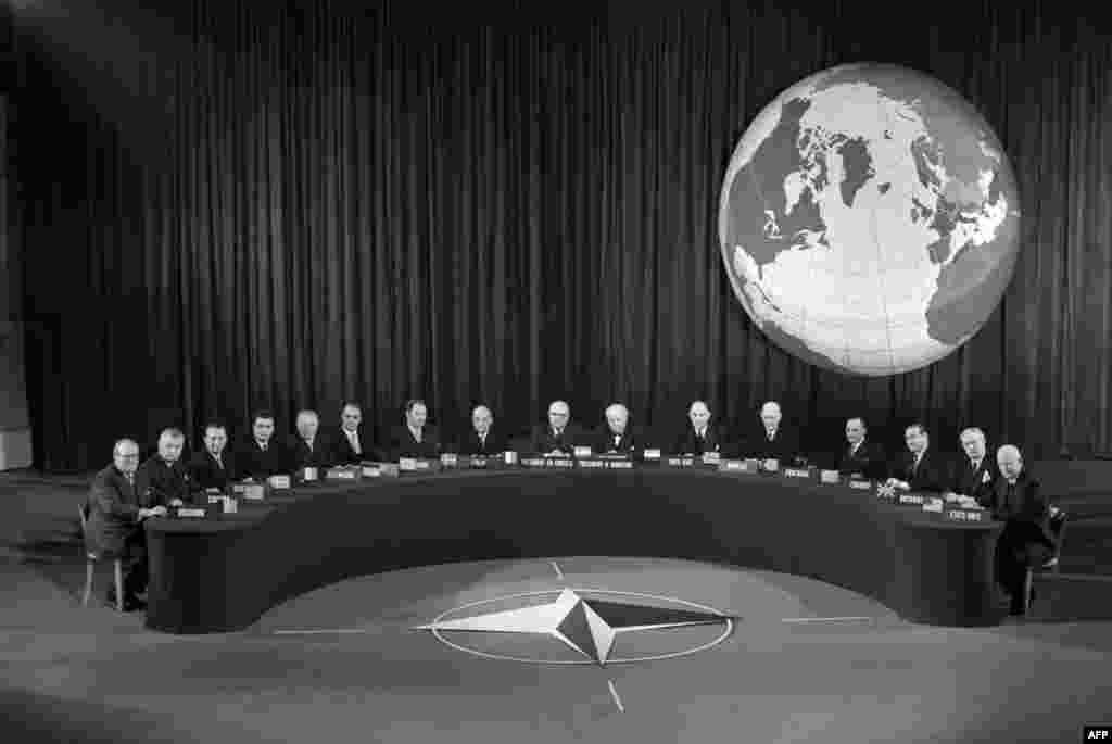 Konferencija NATO-a u Parizu 1957. uz amblem NATO-a koji je usvojen 1953. godine. Zastava NATO-a ima tamnoplavu boju koja predstavlja Atlantski okean, sa kompasom koji simbolizuje smjer prema miru i krugom koji označava jedinstvo.