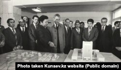 Қазақ Мемлекеттік Университетінің 1970-1986 жылдардағы ректоры Өмірбек Жолдасбеков (қолында таяқша бар) оқу орны макетін Қазақ ССР компартиясының бірінші хатшысы Дінмұхаммед Қонаевқа көрсетіп тұр. Kunaev.kz сайтындағы сурет