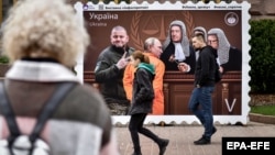 Ljudi prolaze ispred postera koji prikazuje ruskog predsednika Vladimira Putina kojeg kao zarobljenika pred Međunarodni sud u Hagu dovodi ukrajinski general Valerij Zalužnji, Kijev, maj 2023