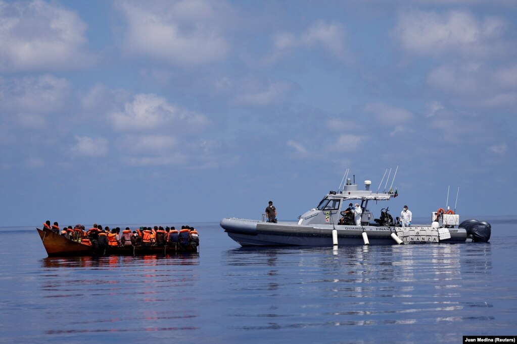 Anëtarët italianë të Guardia di Finanza përgatiten që t'u ndihmojnë migrantëve që janë në varkën prej hekuri.