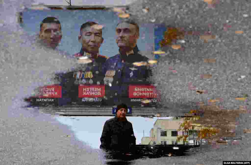 A man walks past a billboard reflecting in a puddle depicting the portraits of Russian Army officers in St. Petersburg.