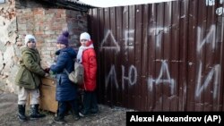 Trei copii stau lângă gardul unei case avariate cu inscripția „Copii și oameni”, în Mariupol, regiunea Donețk controlată de Rusia, sâmbătă, 25 februarie 2023.