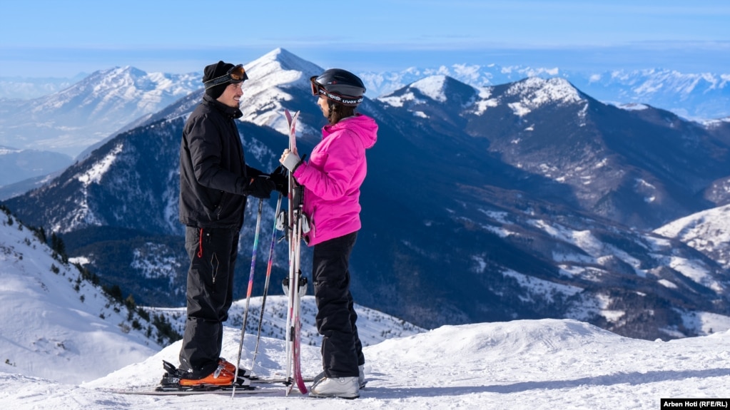 Një djalë dhe një vajzë me ski qëndrojnë pranë njëri-tjetrit ndërsa në sfond shtrihen Malet e Sharrit.