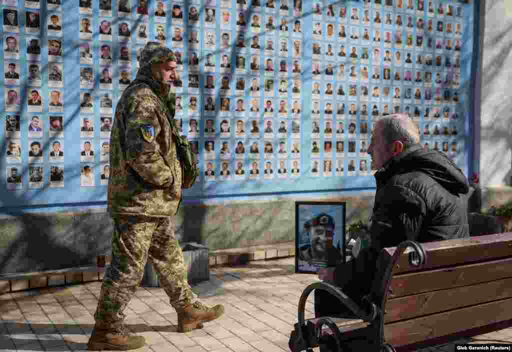 A soldier walks by as Anatoliy, 68, holds a portrait of his son Yuriy 26, who was killed fighting invading Russian forces.