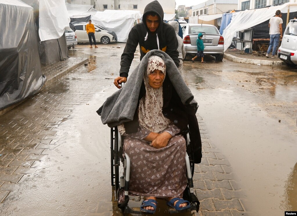Një djalë ndihmon një grua palestineze në një karrocë me rrota në mes të shiut.&nbsp;
