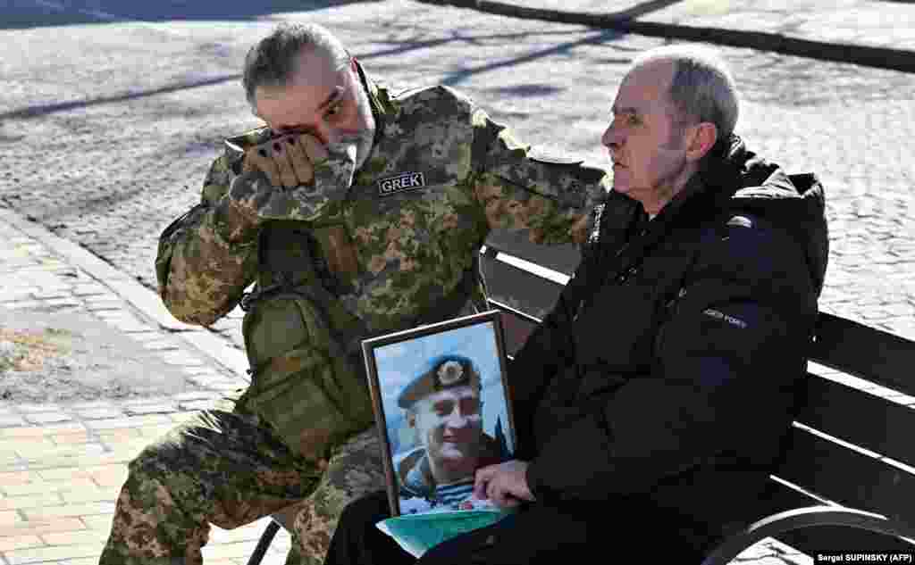 Një ushtar duke fshirë lotët ndërsa qëndron ulur pranë burrit që po mban fotografinë e djalit të tij të rënë.