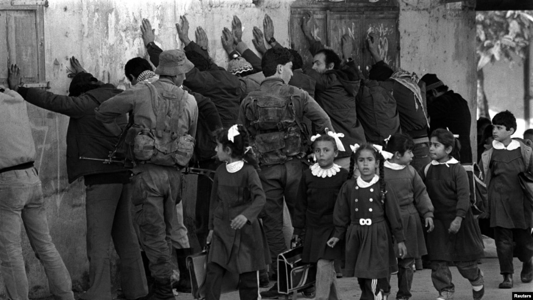 Școlărițe palestiniene care se întorceau acasă de la cursuri trec pe lângă un rând de bărbați arabi percheziționați de soldații israelieni în Fâșia Gaza, în 1986, după ce un evreu a fost înjunghiat și grav rănit. 