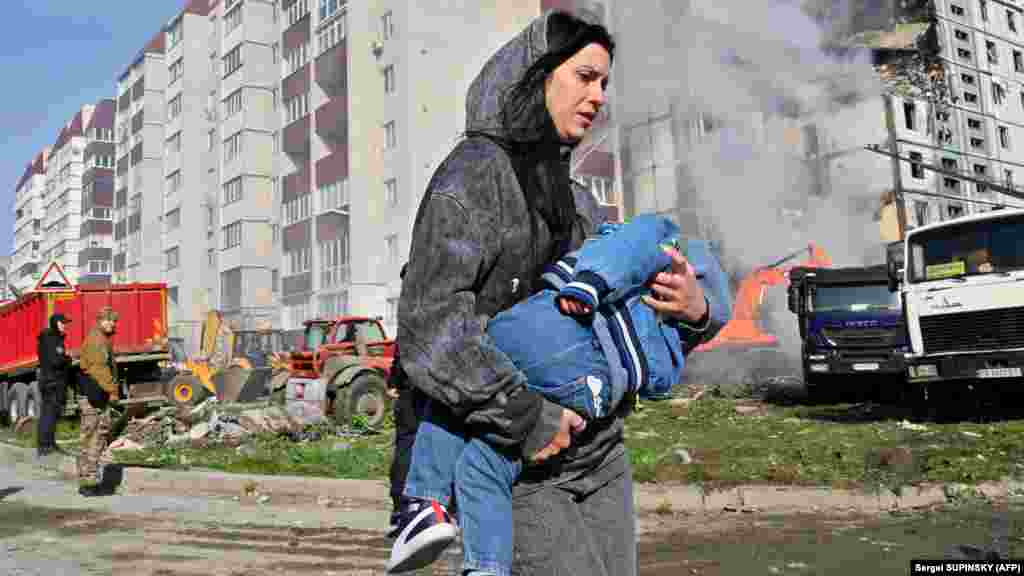 Egy nő karjában viszi gyermekét a füstölgő umani lakóépület mellett az orosz rakétacsapás után