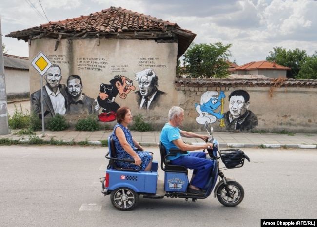Një çift vendas duke kaluar në rrugë ndërsa në një mur shihen grafite figura politike që ndërveprojnë me personazhet e filmave vizatimorë në Staro Zhelezare, Bullgari, më 6 gusht.