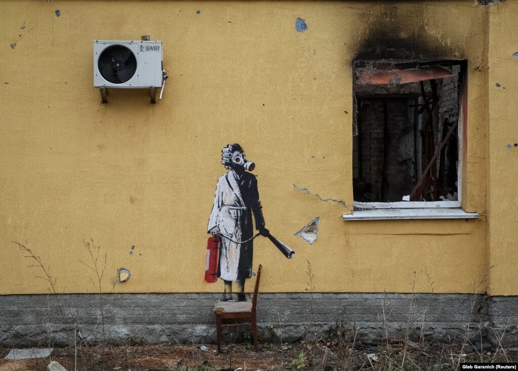 Një grua me robdeshambër, flokët me lidhëse për kaçurrela, e në fytyrë ka të vendosur një maskë kundër gazit, në dorë mban një pajisje për fikjen e zjarrit.