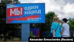 Женщины идут по улице мимо рекламного щита с пророссийскими лозунгами в оккупированном Россией городе Мелитополе Запорожской области, 3 августа 2022 года