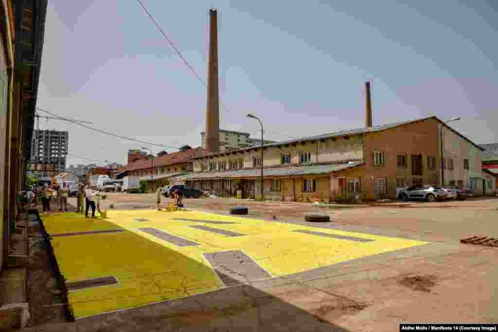 Oamenii pictează o lucrare de artă în aer liber pe asfaltul din afara fostei fabrici de cărămidă din Pristina. Manifesta 14 s-a deschis pe 21 iulie și se va desfășura până pe 30 octombrie. &nbsp;