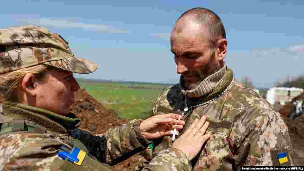 Дружина допомагає чоловікові одягнути на шию вервицю, подаровану військовими капеланами після Великоднього богослужіння на їхній позиції у Харківській області, 24 квітня 2022 року