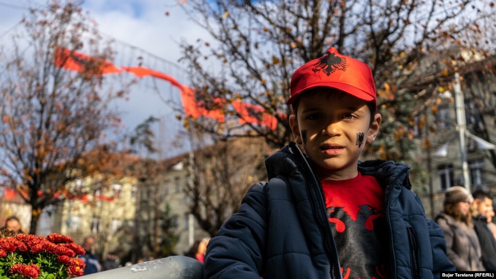 Një djalë i vogël i veshur me bluzë e kapele me ngjyrat e flamurit kombëtar, ndërsa në të dyja faqet ka të pikturuar shqiponjën.