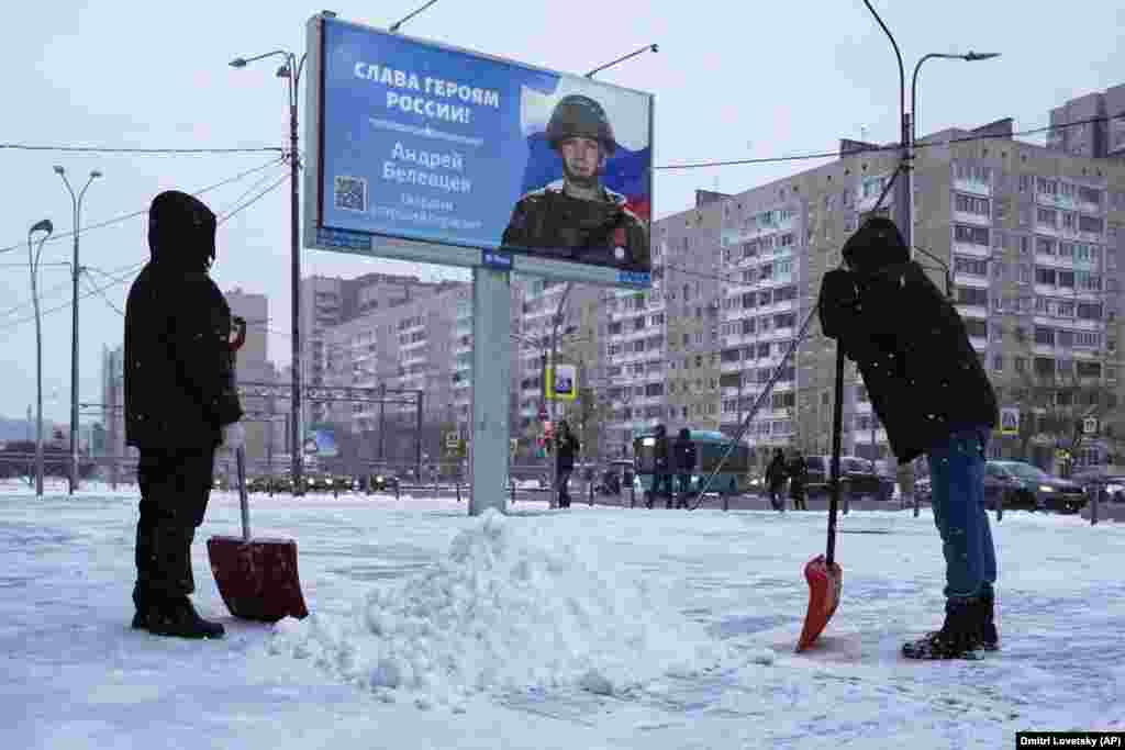 Workers clear snow next to a billboard with a portrait of a Russian soldier and the words &quot;Glory to the heroes of Russia&quot; in St. Petersburg.