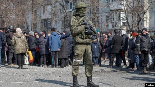 A Russian soldier stands next to local residents waiting in line for humanitarian aid in the besieged southern Ukrainian port of Mariupol. 