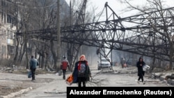 Local residents walk near a fallen electricity pylon and a destroyed apartment building in the besieged southern Ukrainian port city of Mariupol