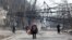 Local residents walk near a fallen electricity pylon and a destroyed apartment building in the besieged southern Ukrainian port city of Mariupol