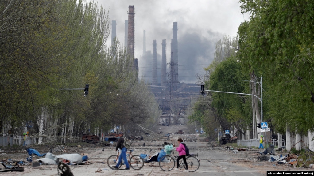 People walk their bikes across a street in Mariupol as smoke rises above the Azovstal steelworks on May 2.