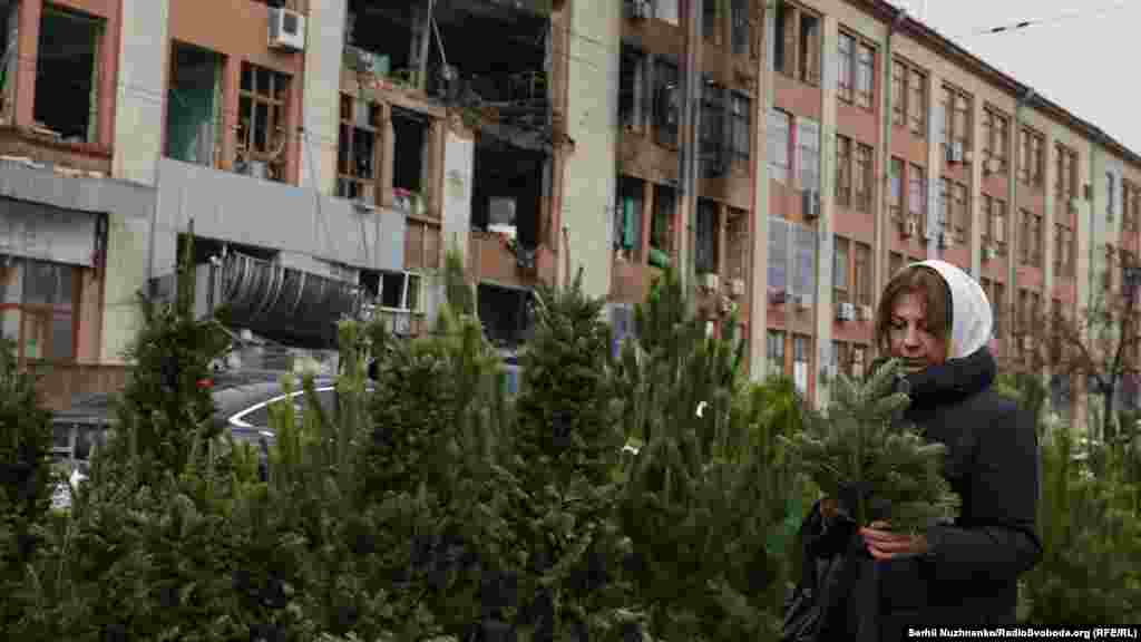 Жінка обирає гілочку ялини перед Різдвом на вуличному ринку неподалік від метро «Лукʼянівська». Позаду неї – зруйнована обстрілами адміністративна будівля