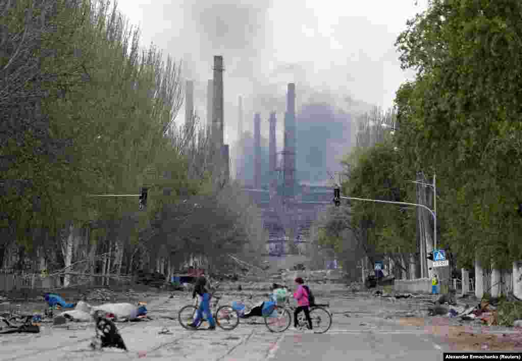 Kerékpárosok sétálnak Mariupol egyik utcáján, a háttérben füst száll fel az Azovstal Vas- és Acélművek egyik üzeméből május 2-án