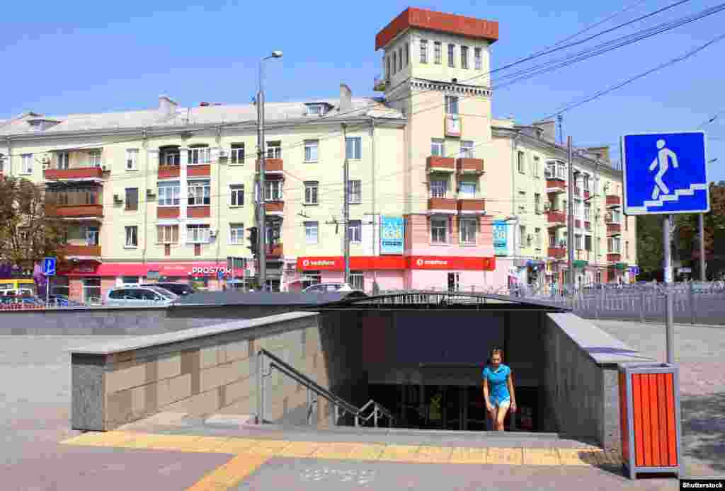 Žena izlazi iz podvožnjaka na raskrsnici u centru Mariupolja. Budući da je ova fotografija snimljena 2019. godine, zgrada u pozadini imala je krov u obliku piramide koji je dodan na kupolu. Na drugoj slici vidi se ista zgrada iz zraka u aprilu 2022.&nbsp;