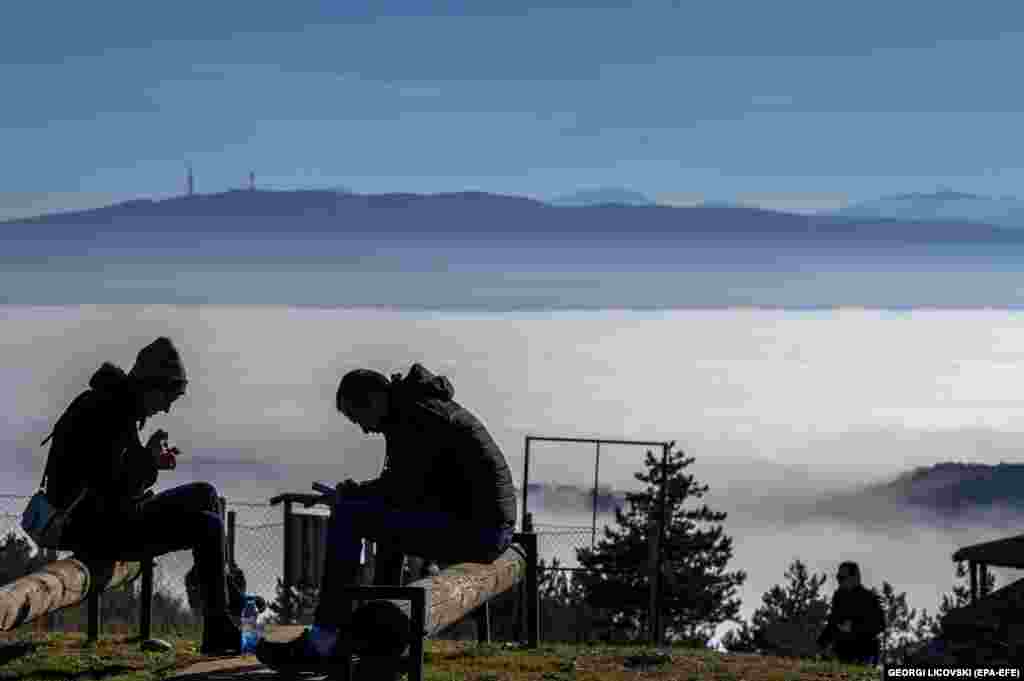 Людзі атрымліваюць асалоду ад сонечнага дня ў Паўночнай Македоніі на гары каля вёскі Кукава, гледзячы на сталіцу Скоп&#39;е, пакрытую смогам.