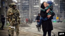 Жена со дете поминува покрај руски војници на улица во Мариупол на 12 април.