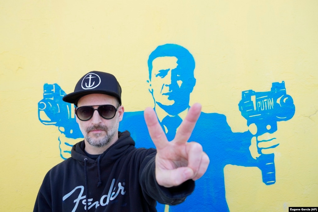 Artisti rrugor, Todd Goodman, i njohur si 1GoodHombre, duke qëndruar para një portreti të presidentit ukrainas, Volodymyr Zelensky, të cilin ai e ka paraqitur duke mbajtur armë zjarri, më 21 mars, në Santa Monika të Kalifornisë, në SHBA. Goodman ka dalë publikisht, duke rrezikuar të arrestohet dhe të gjobitet për veprën e palejuar, për të grumbulluar fonde për refugjatët ukrainas të luftës. Goodman ka thënë për REL-in se veprat e tij janë qëndrime të rezistencës paqësore ndaj agresionit të pakontrolluar të Putinit dhe të dhunës në Ukrainë. &ldquo;Unë nuk e di nëse veprat e mia mund të ndryshojnë diçka fizikisht, përveç se mbase të ndryshojnë ndjenjat e njerëzve, nga ndjenja e pashpresës dhe frikës, në ndjenjën e shpresës dhe trimërisë përballë të keqes&rdquo;.