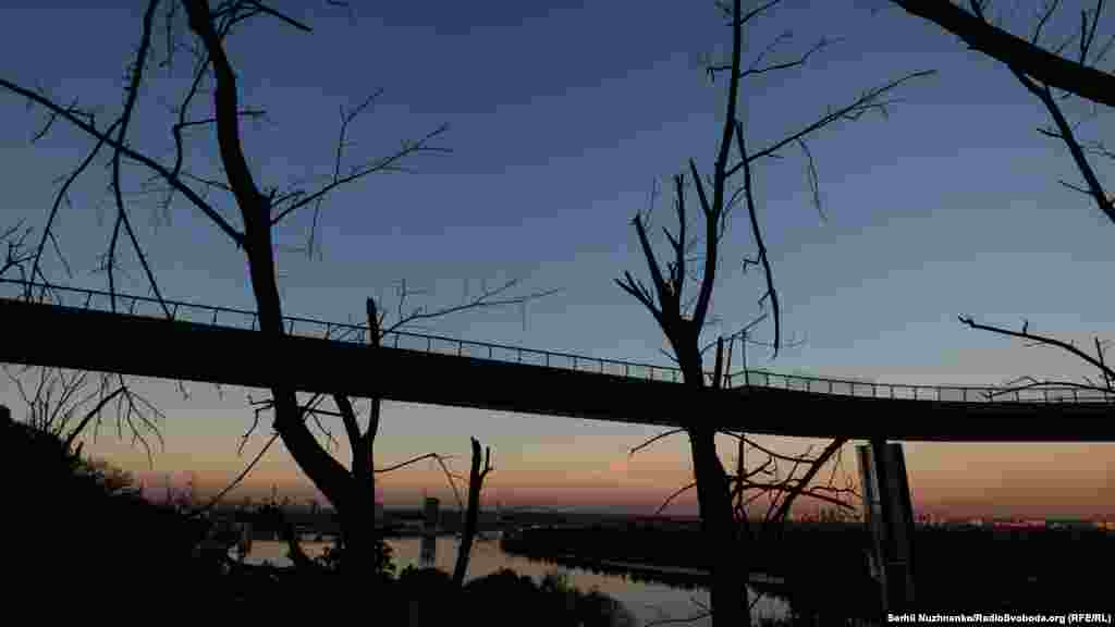 Вечірнє сонце в обрисах обгорілих дерев і пішохідно-велосипедного мосту після влучання поблизу російської ракети