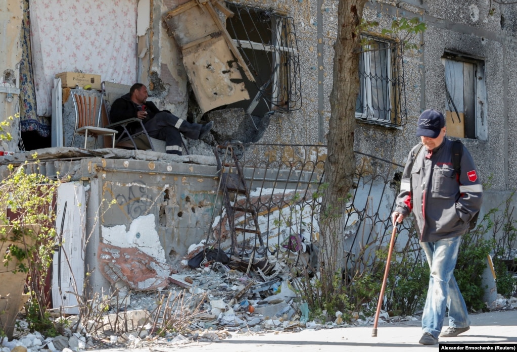 Një burrë duke qëndruar në një karrige para një ndërtese të dëmtuar banimi, më 12 maj. Pasi kanë mbijetuar disa muaj bombardimesh, banorët tani duhet të durojnë pritjen në radhë për ujë dhe ushqim në qytetin e okupuar.