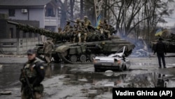 Украинские военнослужащие въезжают на танке в освобожденный от российских войск населенный пункт. На дороге остался разрушенный автомобиль с телом погибшего водителя.