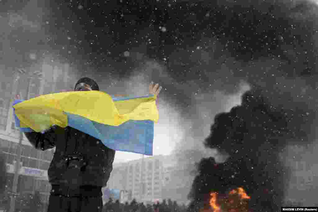 A man holds a Ukrainian flag as smoke rises in the background during clashes between police and pro-European protesters in Kyiv on January 22, 2014.&nbsp;