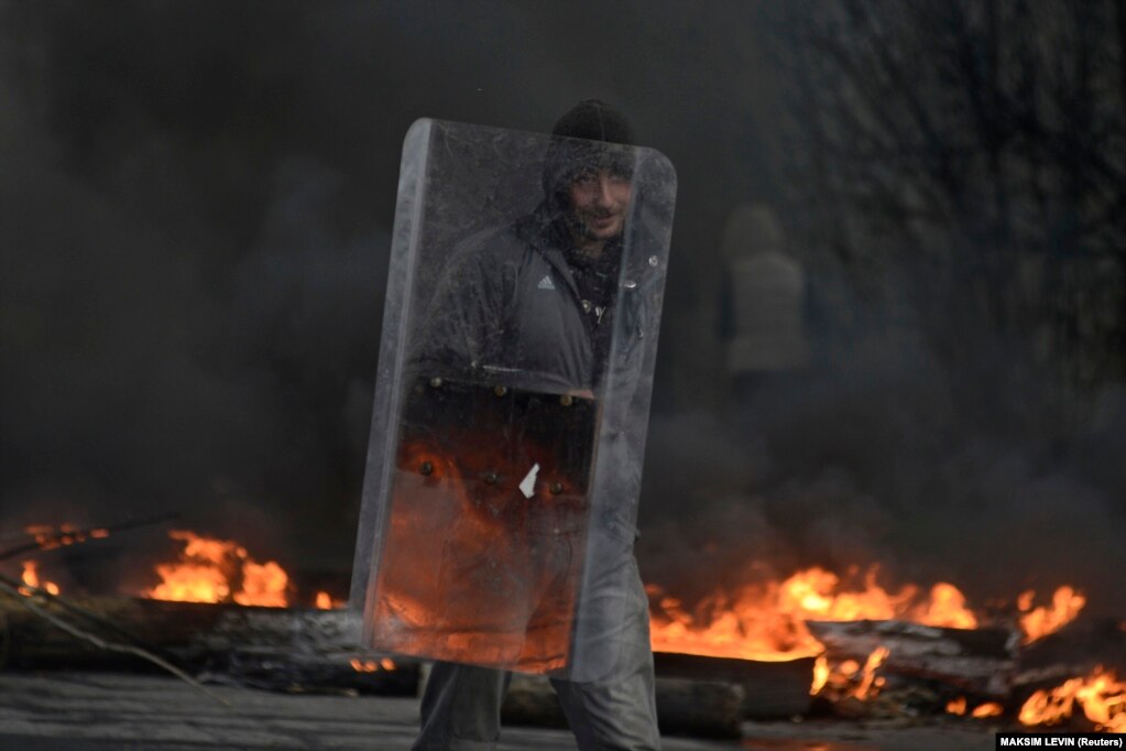 Një protestues pro-Rusisë duke mbajtur një mburojë kundër trazirave në një pikë kontrolli, teksa tymi ngrihej lart nga djegia e gomave, në Sllovjansk, Ukrainë, më 13 prill 2014. Atë vit, ushtria e Rusisë filloi t'i mbështeste forcat separatiste, të cilat, pas betejave me trupat qeveritare ukrainase, morën kontrollin e disa pjesëve të rajoneve të Donjeckut dhe të Luhanskut në Ukrainë.