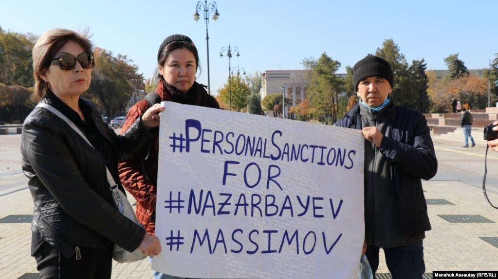 Пришедшие на площадь в Алматы в день анонсированного митинга держат плакат с требованием ввести санкции против экс-президента Назарбаева и главы КНБ Масимова, 23 октября 2021 года