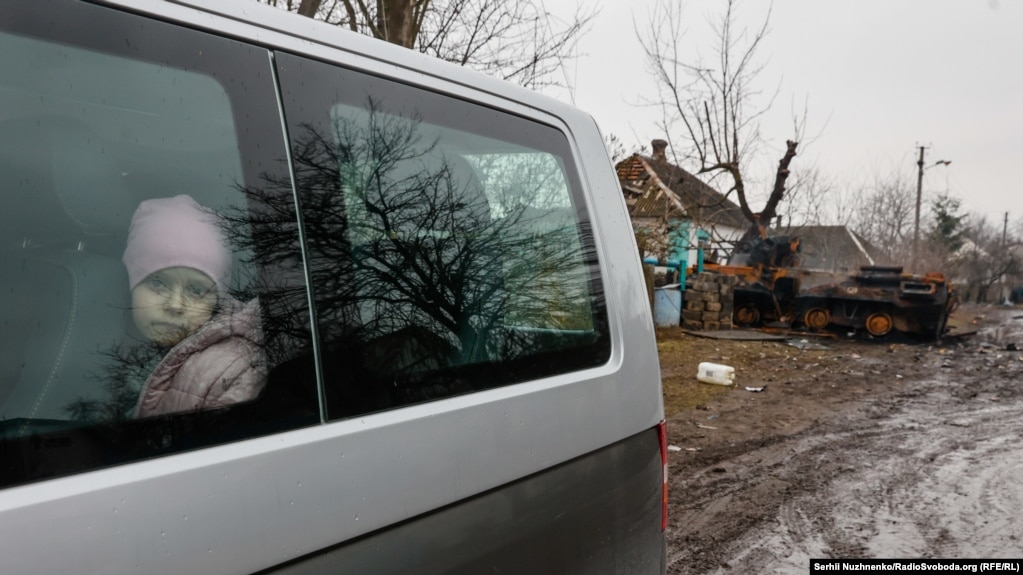 Një fëmijë duke shikuar nga dritarja e një furgoni gjatë evakuimit, ndërsa pas shihet një automjet i blinduar rus që është shkatërruar.