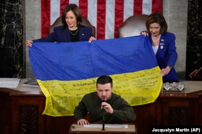 Zëvendëspresidentja e SHBA-së, Kamala Harris (majtas), dhe kryetarja e atëhershme e Dhomës Nancy Pelosi, reagojnë ndërsa presidenti ukrainas, Volodymyr Zelensky, u dhuroi ligjvënësve një flamur ukrainas me autografe nga trupat që po luftojnë në Bahmut. Presidenti ukrainas mbajti një fjalim gjatë një sesioni të përbashkët të Kongresit në Uashington më 21 dhjetor 2022 gjatë vizitës në SHBA.