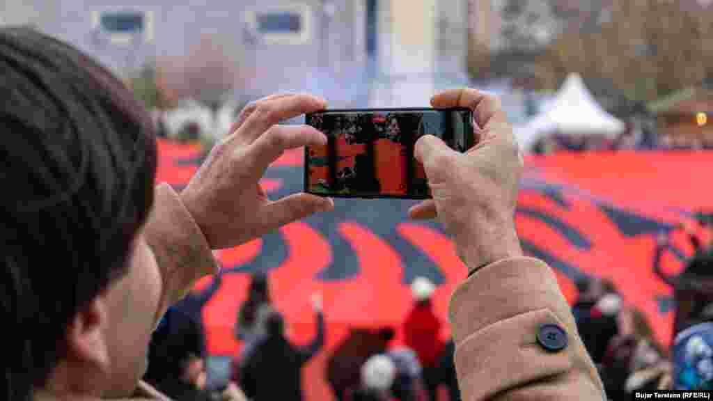 Një burrë duke regjistruar me telefonin e tij shpalosjen e një flamuri me përmasa të mëdha në mes të sheshit të Prishtinës.