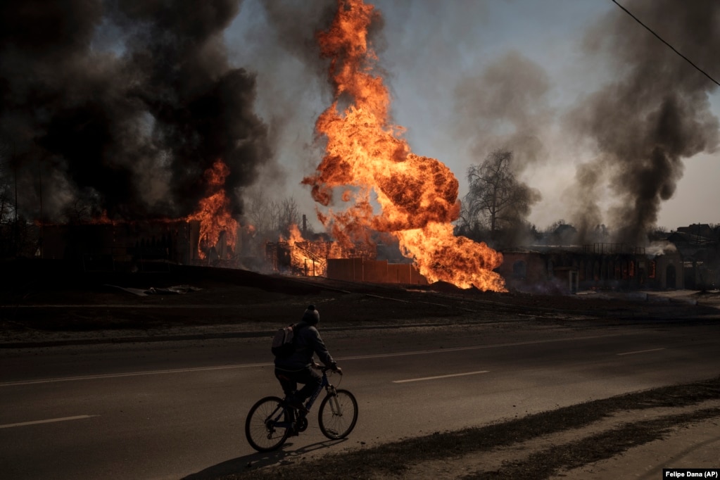 Një burrë duke kaluar me biçikletë jo larg tij shihen flakët dhe tymi që dalin nga zjarri pas një sulmi rus në Harkiv më 25 mars 2022.