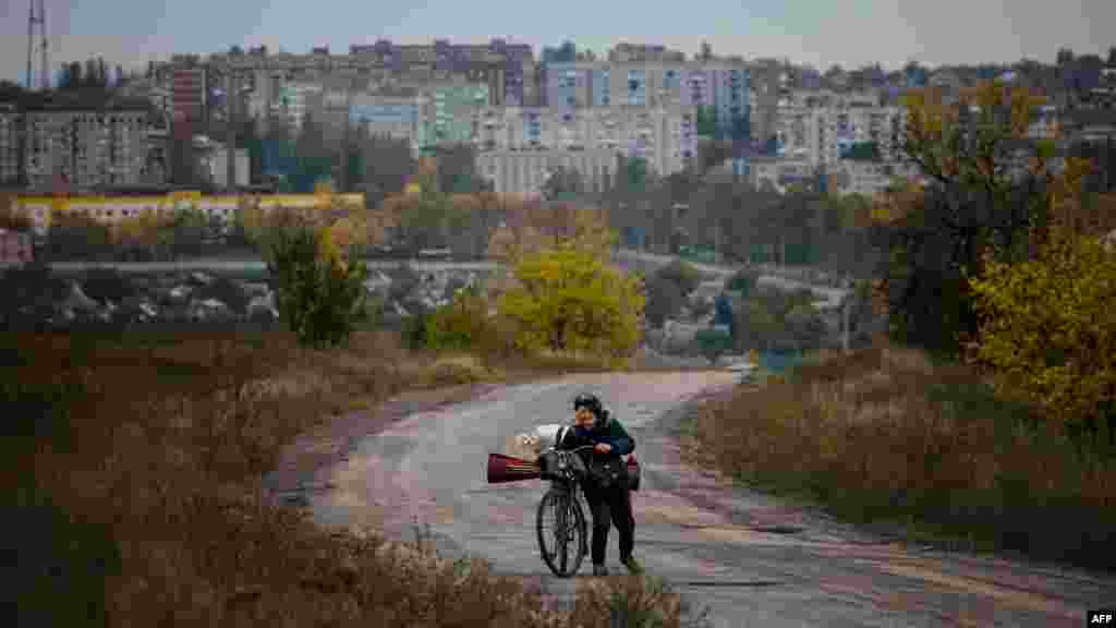 Egy idős asszony tolja holmijával megrakott kerékpárját, mögötte Bahmut látképe