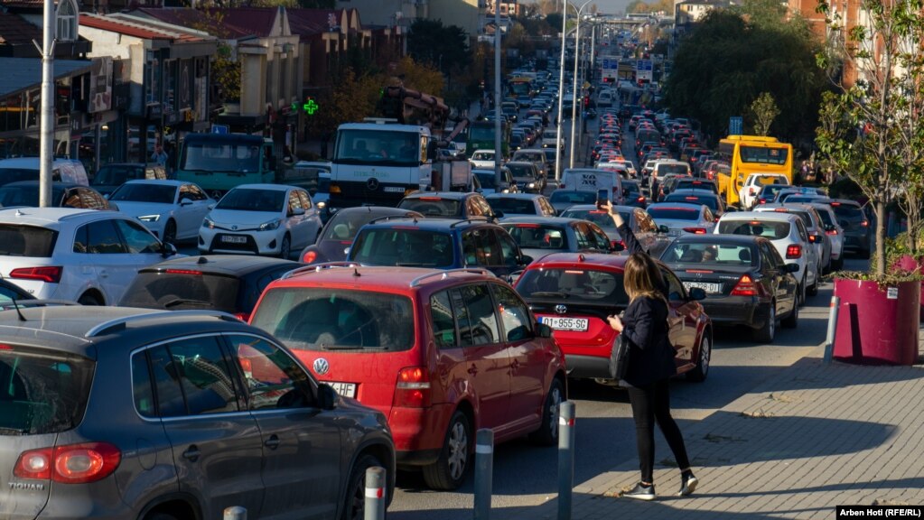 Një këmbësore duke përshëndetur dikë ndërsa shihet trafiku i mbingarkuar me automjete.